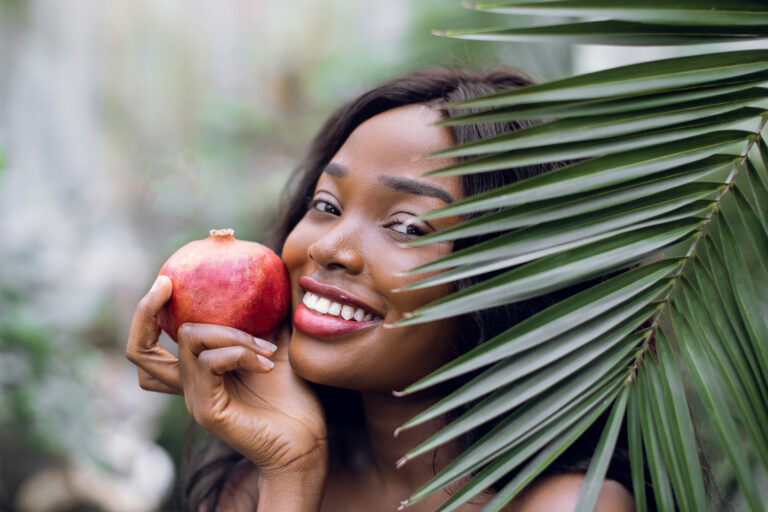 Close up portrait of young emotional beautiful afro american woman with pomegranate in her hands, looking out of green plam leaf indoors in greenhouse or garden. summer and beauty concept